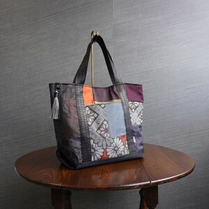 以下が含まれることがあります： 紫、オレンジ、グレー、青の色合いの様々な柄の生地を使ったパッチワークトートバッグ。バッグには黒いハンドルと黒いフェイクレザーのトリムが付いています。グレーのタッセルが側面からぶら下がっています。バッグは丸い木製のテーブルの上にあります。