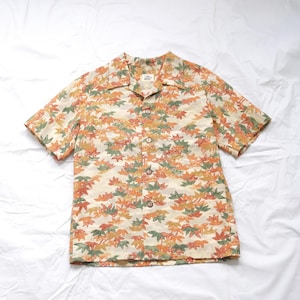 May include: Short-sleeved button-down shirt with a cream-colored background and a repeating pattern of orange, green, and yellow leaves. The shirt has a collared neckline and wooden buttons. The shirt is made of a lightweight fabric.