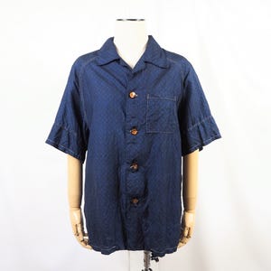 Puede incluir: Una camisa de manga corta con botones en tela azul oscuro tipo mezclilla con un sutil patrón geométrico. La camisa presenta un cuello clásico, un bolsillo en el pecho en el lado izquierdo y cuatro botones marrones jaspeados en la parte frontal.