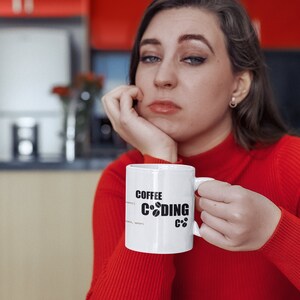 K&ouml;nnte beinhalten: Eine wei&szlig;e Kaffeetasse mit schwarzem Text, der "Coffee Coding C++" lautet, und einem Kaffeekorn-Symbol. Die Tasse wird von einer Person gehalten, die einen roten Pullover tr&auml;gt.