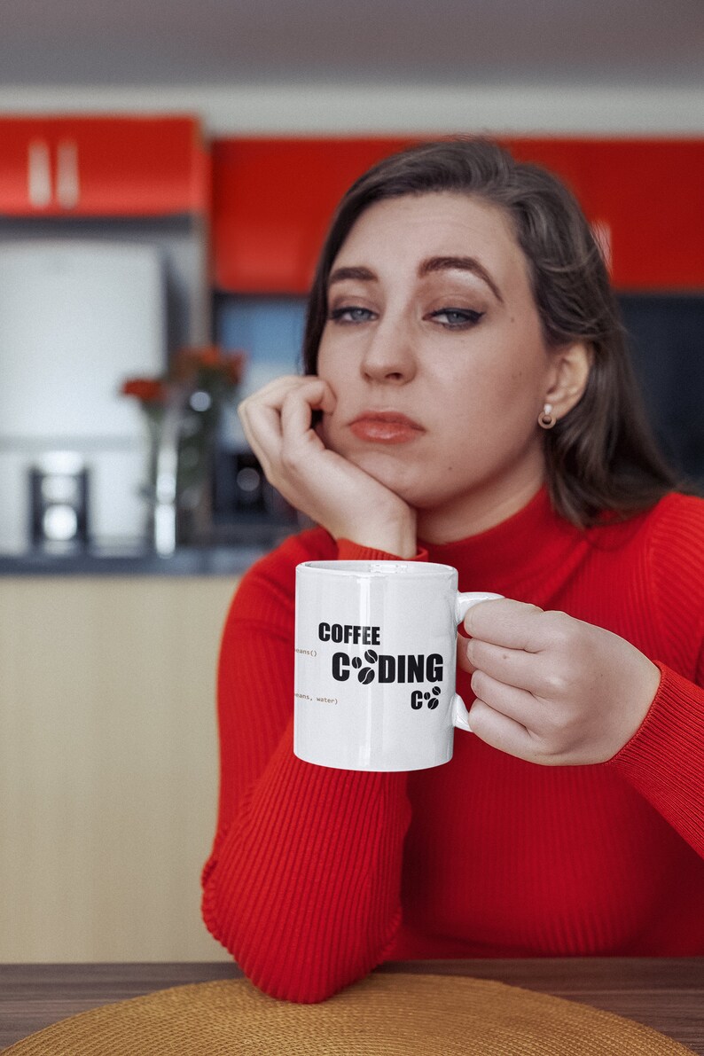 K&ouml;nnte beinhalten: Eine wei&szlig;e Kaffeetasse mit schwarzem Text, der "Coffee Coding C++" lautet, und einem Kaffeekorn-Symbol. Die Tasse wird von einer Person gehalten, die einen roten Pullover tr&auml;gt.