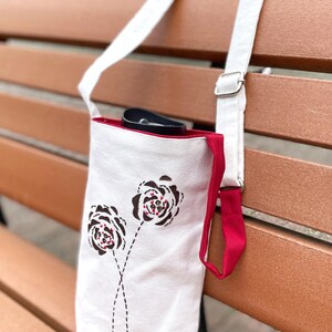 May include: White canvas tote bag with a red interior and strap. The bag features two stylized brown and red floral designs. The bag has a white adjustable strap and a red accent strap. A black object is visible inside the bag.