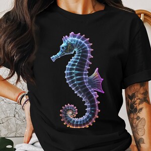 以下が含まれることがあります： カラフルな虹色のタツノオトシゴのグラフィックがプリントされた黒いTシャツ。タツノオトシゴは左を向いており、青、紫、ピンクの色合いです。