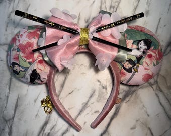 May include: Pink mouse ears headband with a large pink bow, adorned with white flower accents and black chopsticks. The ears feature a floral print with a cartoon character. A gold charm hangs from the headband.
