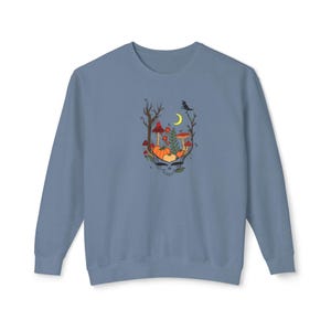 May include: A light blue sweatshirt featuring a colorful graphic design. The design includes a skull with pumpkins, mushrooms, and a crescent moon. A crow sits on a branch. The sweatshirt is made of a soft material.