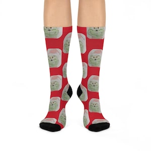 May include: Red knee-high socks featuring a repeating pattern of a green, cartoonish face wearing a Santa hat. The socks have black heels and toes. The design is a festive, holiday-themed pattern.