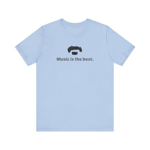 May include: A light blue short-sleeve t-shirt featuring a black graphic of a mustache centered above the text "Music is the best." in a dark gray sans-serif font. The garment has a crew neckline and a standard fit with short sleeves.
