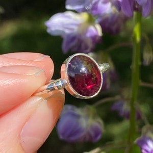 May include: A silver ring featuring a deep red gemstone set in a bezel setting. The ring is worn on a finger, with a blurred background.