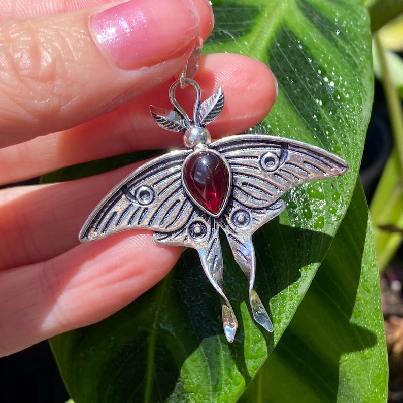 May include: A silver-toned moth pendant with a large, teardrop-shaped, deep red gemstone at its center. The pendant features detailed wing patterns and delicate antennae. The jewelry is held against a backdrop of green foliage.