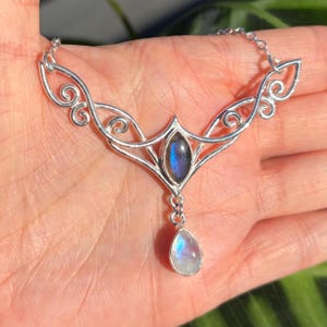 May include: A silver-toned necklace featuring a central marquise-shaped labradorite gemstone with blue iridescence, accented by a dangling teardrop-shaped moonstone, set within an intricate, swirling metal frame attached to a delicate chain.