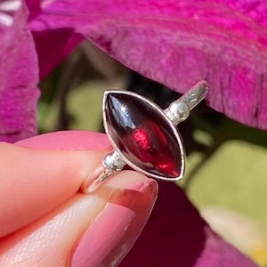 May include: A silver ring with a marquise-shaped, deep red gemstone. The ring has a thin band with small silver decorative accents. The gemstone is set in a silver bezel. The ring is shown against a blurred background of pink and green. The ring's design is elegant and simple.
