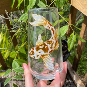 May include: A clear glass with a hand-painted design of a white and orange koi fish. The fish is swimming upwards with its tail fin extended.