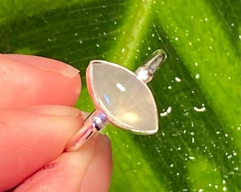May include: A silver ring featuring a marquise-cut, pale, iridescent gemstone. The ring is set against a green backdrop, accentuating the gemstone's subtle hues and the ring's polished metal.