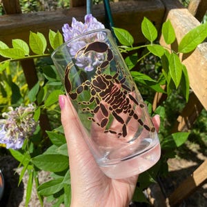 Può includere: Un bicchiere di vetro trasparente con un design di scorpione dorato e nero. Il bicchiere è tenuto da una mano.