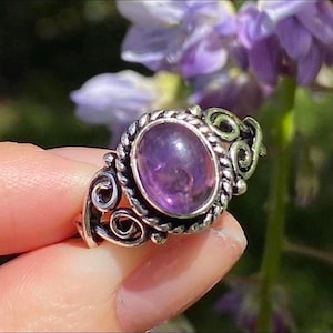 May include: A silver ring featuring an oval, deep purple amethyst gemstone. The ring has an ornate, swirling design around the gemstone and along the band. The ring is held up in front of a blurred background of purple flowers.