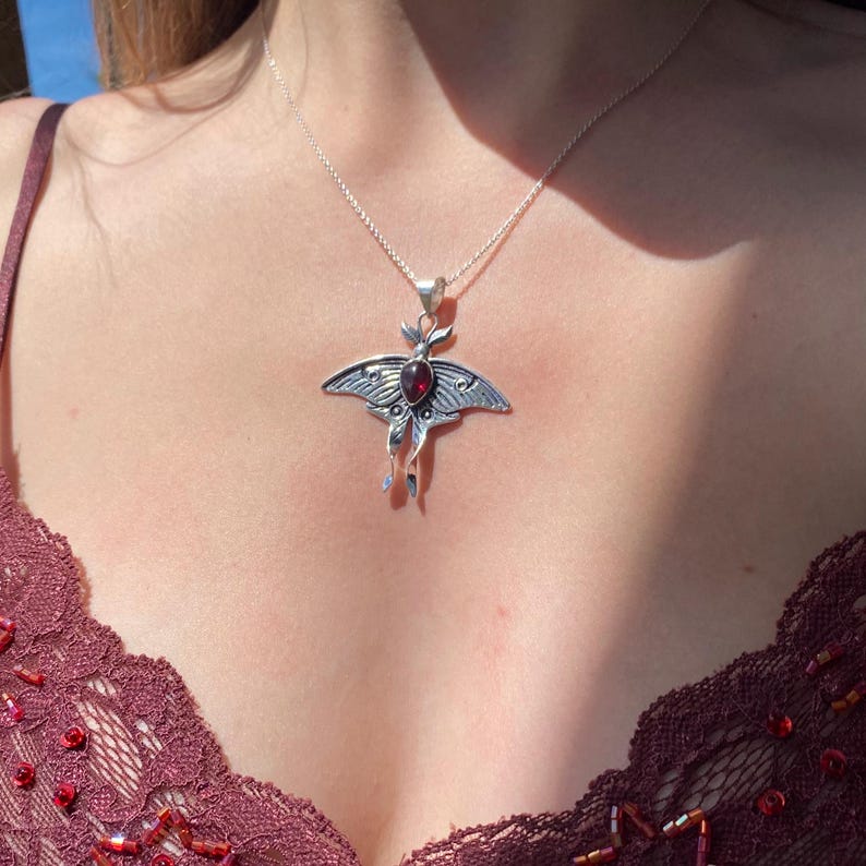 May include: A silver-toned pendant necklace featuring a moth design with a central red gemstone. The moth pendant has detailed wing patterns and delicate antennae. The necklace is worn on a silver chain, creating a striking contrast against the skin.