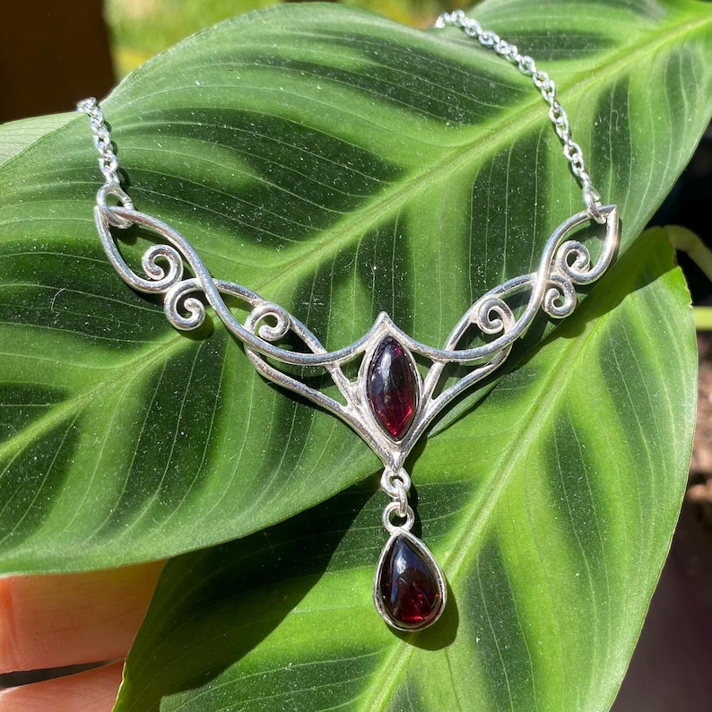May include: A silver-toned necklace with a decorative openwork design, featuring two oval-shaped, deep red gemstones and a teardrop-shaped pendant. The necklace is displayed on a large green leaf.