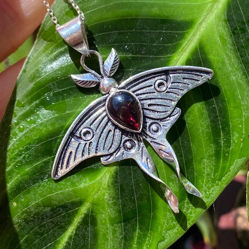 May include: A silver-toned moth pendant necklace with a teardrop-shaped, dark red gemstone at its center. The moth's wings have detailed etched patterns. The necklace is displayed on a large, green leaf.