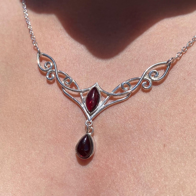 May include: A silver-toned necklace with a garnet pendant. The necklace features an ornate design with a central marquise-shaped garnet and a teardrop-shaped garnet pendant. The chain is delicate and the necklace is worn around the neck.
