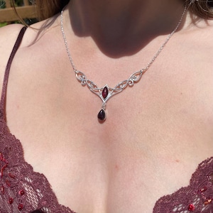 May include: A silver necklace with a garnet pendant. The necklace features an ornate design with a central marquise-cut garnet and a teardrop-shaped garnet pendant. The necklace is worn on a person's chest, with a burgundy lace top visible.