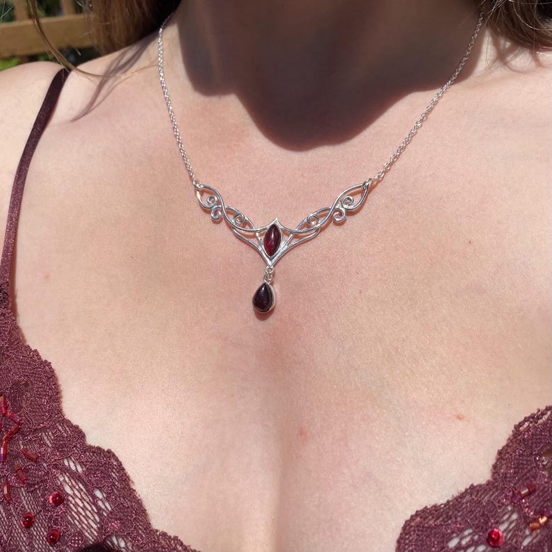 May include: A silver necklace with a garnet pendant. The necklace features an ornate design with a central marquise-cut garnet and a teardrop-shaped garnet pendant. The necklace is worn on a person's chest, with a burgundy lace top visible.