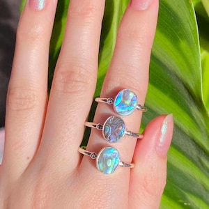 May include: Three silver rings with iridescent abalone shell inlays. The rings are stacked on a finger, with the largest ring on top and the smallest ring on the bottom.