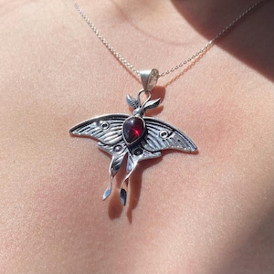 May include: A silver-toned moth pendant necklace. The pendant features a detailed moth design with a central, oval-shaped, red gemstone. The moth's wings are intricately patterned, and the necklace has a delicate chain.