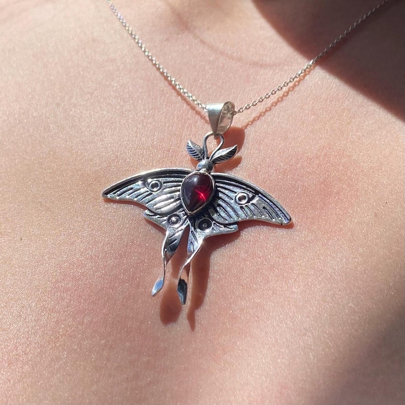 May include: A silver-toned moth pendant necklace. The pendant features a detailed moth design with a central, oval-shaped, red gemstone. The moth's wings are intricately patterned, and the necklace has a delicate chain.