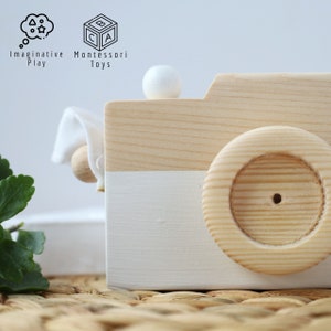 May include: A wooden toy camera with a white painted bottom half and a natural wood top half. The camera has a white knob on top and a wooden wheel on the side.