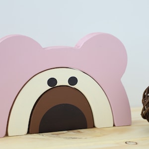 May include: A pink, cream and brown wooden rainbow arch with a bear face. The arch is made of three interlocking pieces.