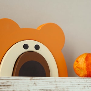 May include: Orange, brown, and cream wooden toy shaped like a bear with a large open mouth. The toy is on a whitewashed wooden shelf with a small orange pumpkin to the right.