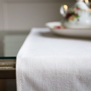 May include: A white table runner with a textured surface. The runner is on a glass table with a gold trim.