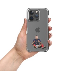 Puede incluir: Una funda de teléfono transparente con la parte posterior gris y una imagen de un jugador de fútbol que lleva una camiseta azul y blanca con pantalones cortos rojos. El jugador está sentado con las piernas cruzadas y los brazos cruzados. La funda tiene un logotipo de Apple negro en la parte posterior.