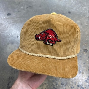 May include: A tan corduroy baseball cap featuring a red embroidered hog mascot with the text HOGS in white. The cap includes a decorative cream-coloured braided rope trim across the base of the crown and a matching tan button on the top centre.