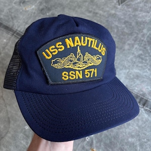 Puede incluir: Gorra de camionero azul marino con parte trasera de malla y visera curva. La parte delantera presenta un parche con el texto "USS NAUTILUS" y "SSN 571" en letras doradas, junto con un emblema dorado.