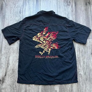 May include: Black short-sleeved button-up shirt with a red, yellow and orange eagle graphic on the back. The eagle is surrounded by flames and the text "Harley-Davidson" is printed below it.