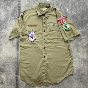 May include: An olive green short-sleeve button-down shirt featuring various embroidered patches, including a Boy Scouts of America emblem, a Quapaw Area Council patch, a Batesville Ark 151 patch, and a small American flag on the right sleeve.