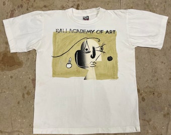 May include: White t-shirt with a graphic print of a stylized face and the text "Bali Academy of Art".