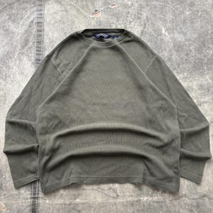 Pode incluir: Uma camisola de manga comprida em verde azeitona com decote redondo e mangas raglan. A peça apresenta uma etiqueta interior no pescoço em azul escuro com o texto branco Northtree Stone e uma pequena etiqueta de tamanho por baixo.