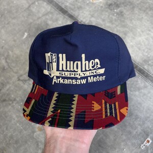 Puede incluir: Una gorra de béisbol azul marino con visera estampada. La gorra presenta el texto "Hughes SUPPLY, INC. Arkansaw Meter" en blanco. La visera tiene un diseño geométrico en rojo, dorado, verde y negro.