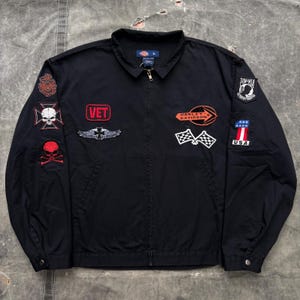 May include: A black Dickies brand zip-up jacket featuring various embroidered patches. The patches include a skull, a cross, the text VET, a Harley-Davidson logo, checkered flags, a POW-MIA emblem, and a USA flag with the number one.
