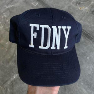 Op de afbeelding: Marineblauwe baseballcap met de wit geborduurde letters "FDNY" op de voorkant. De cap heeft een gebogen klep en een gestructureerde kroon. Een klassieke stijl.