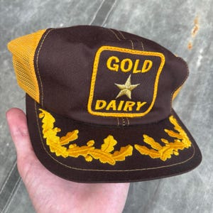 May include: A brown and yellow trucker cap with the words "GOLD DAIRY" embroidered on the front. The cap has a gold star and leaf design. The back of the cap is mesh and yellow.