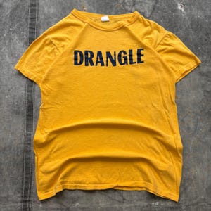 May include: A short-sleeved crew neck t-shirt in a vibrant mustard yellow color. The front of the garment features the word DRANGLE printed in bold, dark navy blue capital letters across the chest area. The shirt has a standard fit and hem.
