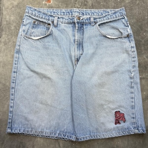 Pode incluir: Calções de ganga azul claro com um design clássico de cinco bolsos. Apresentam um patch de bulldog vermelho e castanho na perna inferior esquerda. Os calções têm fecho de botão e zíper e presilhas para cinto. O denim tem uma aparência desbotada.