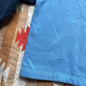 May include: A close-up of a light blue t-shirt with a folded hem. The t-shirt is lying on a brown and white patterned surface.