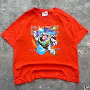 Op de afbeelding: Een oranje t-shirt met een afbeelding van Buzz Lightyear uit Disney's Toy Story. Het ontwerp bevat het personage in zijn ruimtepak, de woorden "BUZZ LIGHTYEAR" en het Disney-logo.