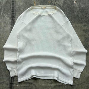 May include: A white, long-sleeved thermal shirt with a waffle-knit texture. The shirt has a crew neck and ribbed cuffs. The fabric appears to be a soft, comfortable material. The shirt is laid flat on a neutral surface.