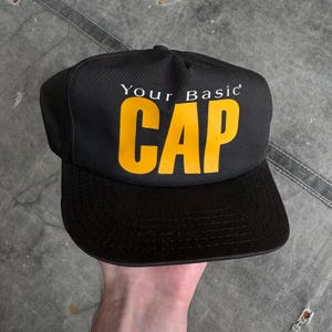 May include: Black baseball cap with the text "Your Basic CAP" in yellow block letters. The cap has a curved bill and a structured crown. The hat is a classic style, suitable for casual wear.