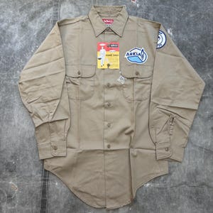 Könnte beinhalten: Beige Arbeitshemd mit langen Ärmeln, Knopfleiste und zwei Brusttaschen. Das Hemd hat Aufnäher mit dem "ARKLA"-Logo und einem runden Emblem auf dem Ärmel. Ein Etikett mit einer Grafik einer Person in Arbeitskleidung ist angebracht.